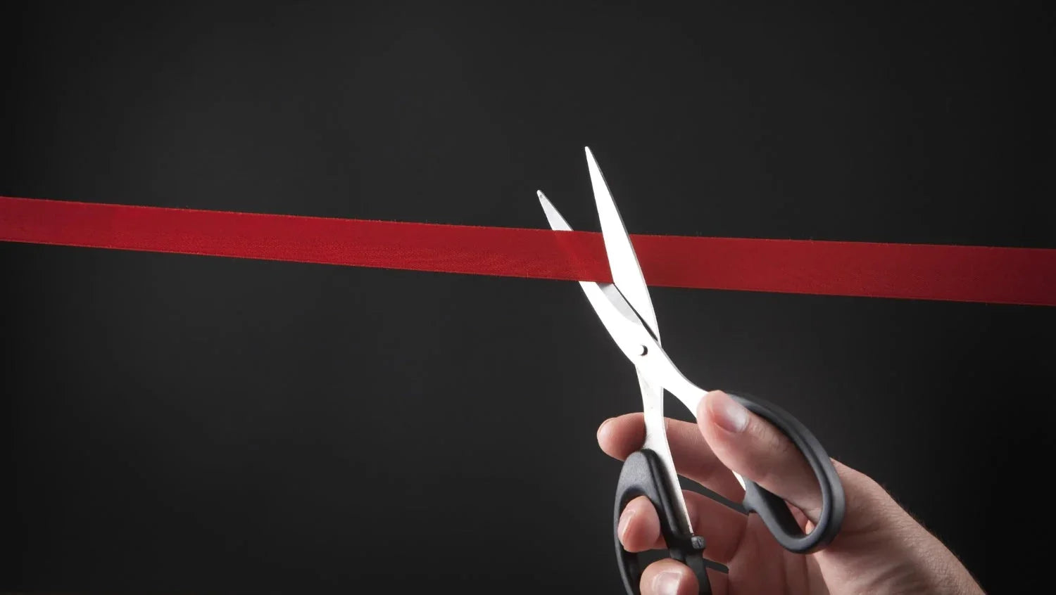 A hand using silver scissors to cut a red ribbon against a dark background.