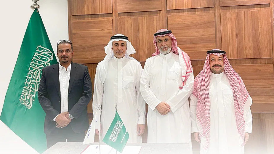Four professional men in Saudi attire and business suit standing before a Saudi Arabian flag.