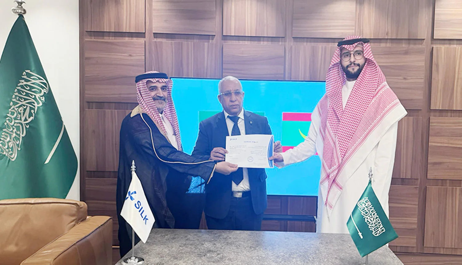 Three men in formal attire holding a certificate in an office with Saudi flags.